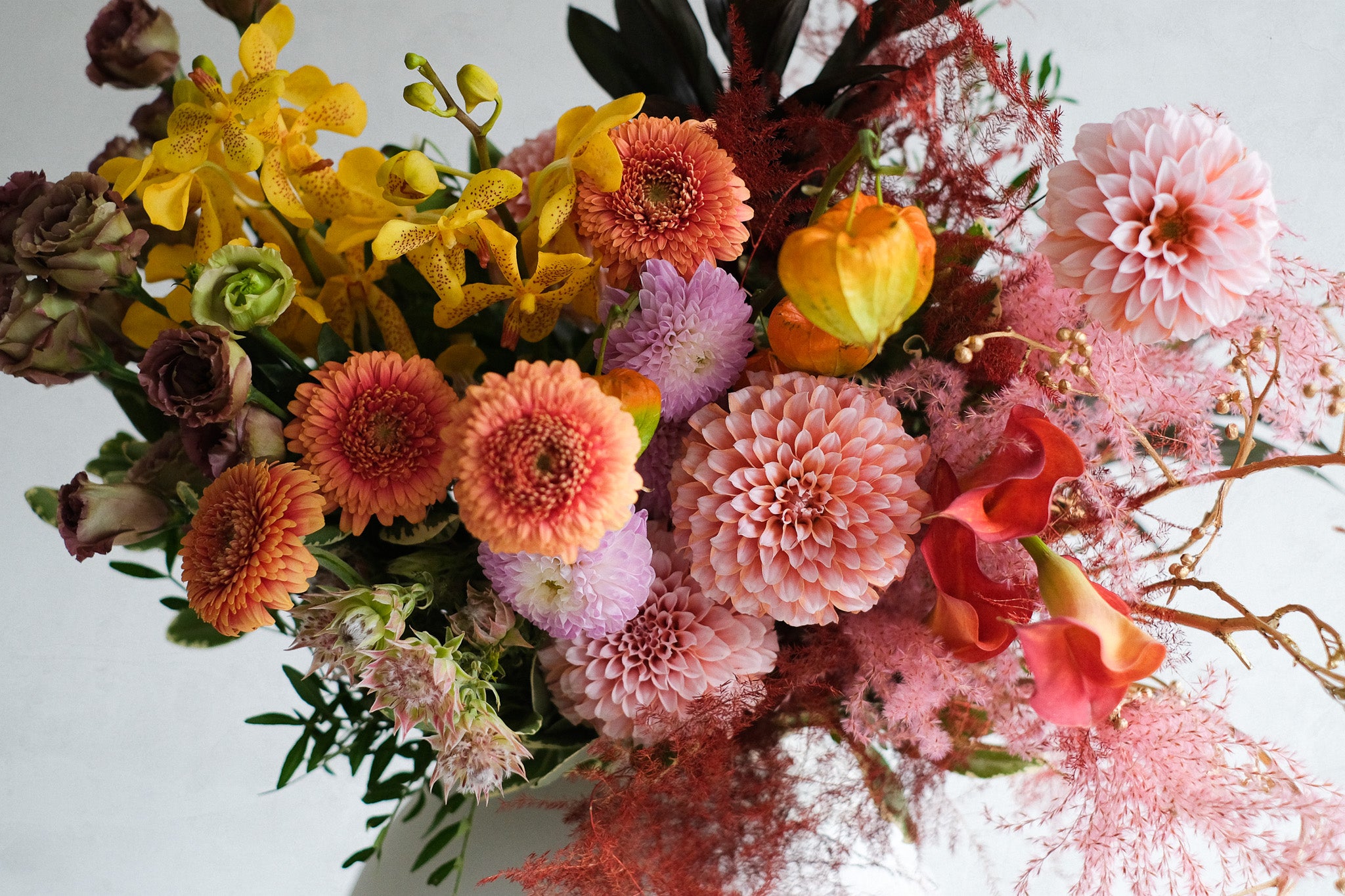 A vibrant floral arrangement featuring a mix of orange, pink, yellow, and purple flowers with green foliage, photographed from a high angle against a plain white background.
